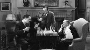 Three men are engaged in a chess game in a vintage room. One man holds a pipe, another watches intently, and the third, with a bandaged arm, looks on from an armchair.