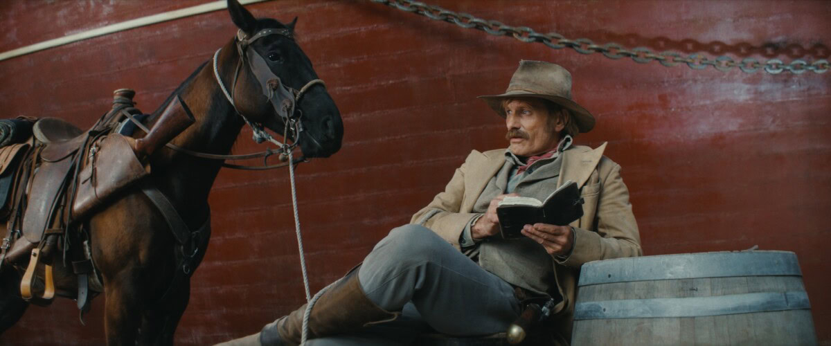 A person in Western attire sits on a barrel reading a book, with a saddled horse standing beside them in front of a red wall.