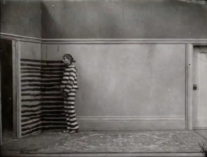 A black and white still of a person in a striped outfit peeking through partially closed vertical blinds in a vintage room.