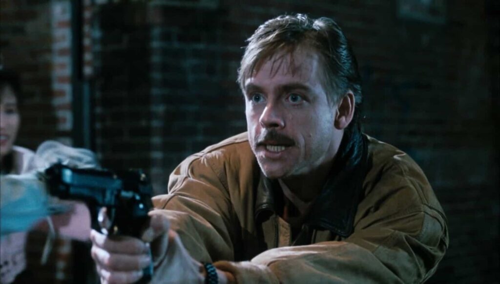 A man with a mustache in a brown jacket aims a handgun, with a brick wall in the background. Another person is partially visible on the left.