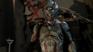 Person in a detailed, alien-like costume with glowing elements stands in an industrial setting; graffiti on the wall behind them reads "Free".
