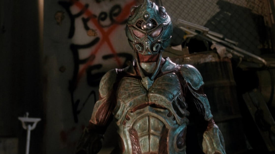 Person in a detailed, alien-like costume with glowing elements stands in an industrial setting; graffiti on the wall behind them reads "Free".