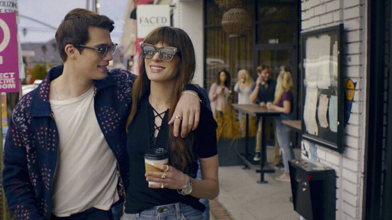 ‘The Idea Of You’ Review – Anne Hathaway And Nicholas Galitzine Sparkle In Their Charming, Thoughtful Rom-Com A young couple, the man wearing a leather jacket and the woman in a black top, walk arm in arm, smiling, with a coffee shop and other people in the background.