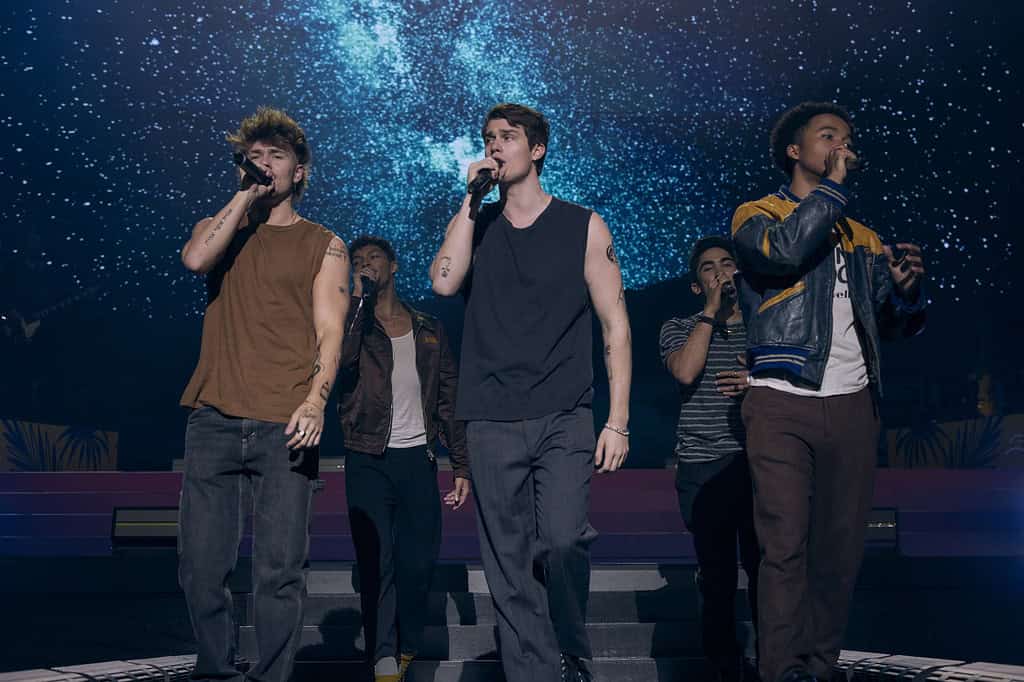 Three young men singing on stage, with a cosmic blue backdrop, accompanied by two other vocalists in the background.
