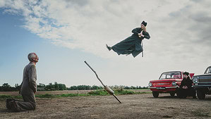 ‘The Invisible Fight’ Blu-Ray Review – A Rock-And-Roll Martial Arts Journey To Enlightenment A man in traditional attire is suspended mid-air by a wooden plank, watched by an older man kneeling and others by a red car in a rural setting.