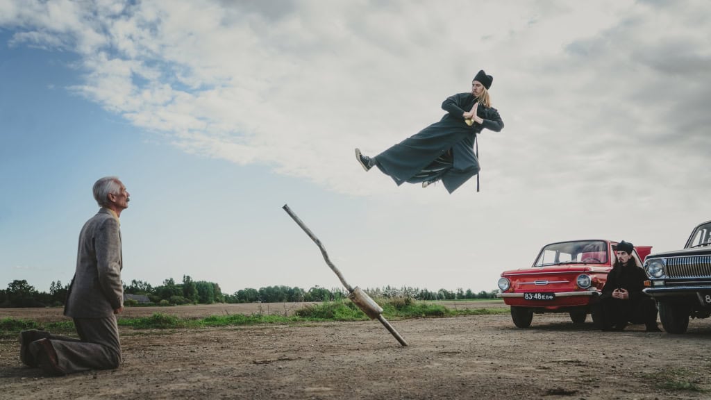 A man in traditional attire is suspended mid-air by a wooden plank, watched by an older man kneeling and others by a red car in a rural setting.
