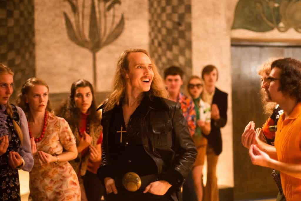 A man in a black leather jacket and large belt buckle stands confidently in a room with retro-dressed people, under warm lighting.