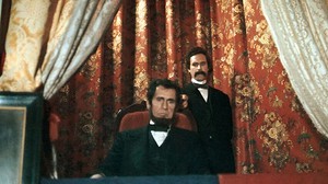 ‘The Lincoln Conspiracy’ Blu-Ray Review – A Passably Entertaining Bit Of Speculative Fiction A still from a film showing an actor portraying abraham lincoln sitting solemnly in a theater box with another actor standing behind him, both in period costumes.