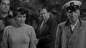 Four people, three men and one woman, standing together with serious expressions in a black and white film scene.