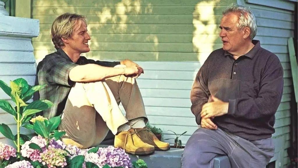 Two men sitting and chatting on a porch; one younger with blond hair and casual clothes, the other older in a sweater, against a backdrop of greenery and flowers.