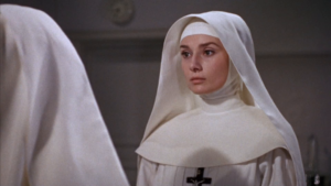 A nun in a white habit with a serious expression stands indoors, facing another person.