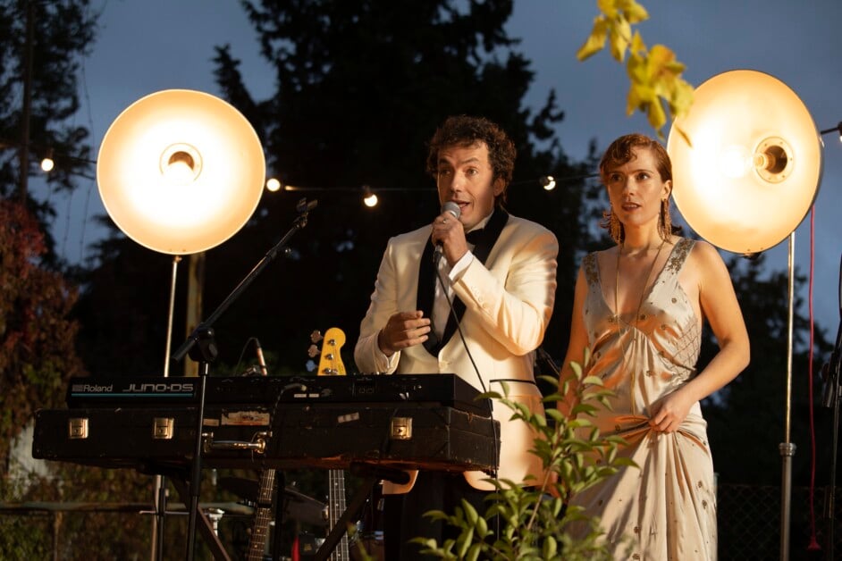 A man in a white suit and a woman in a silver dress stand together on an outdoor stage with microphones. Musical instruments and bright stage lights are visible in the background.