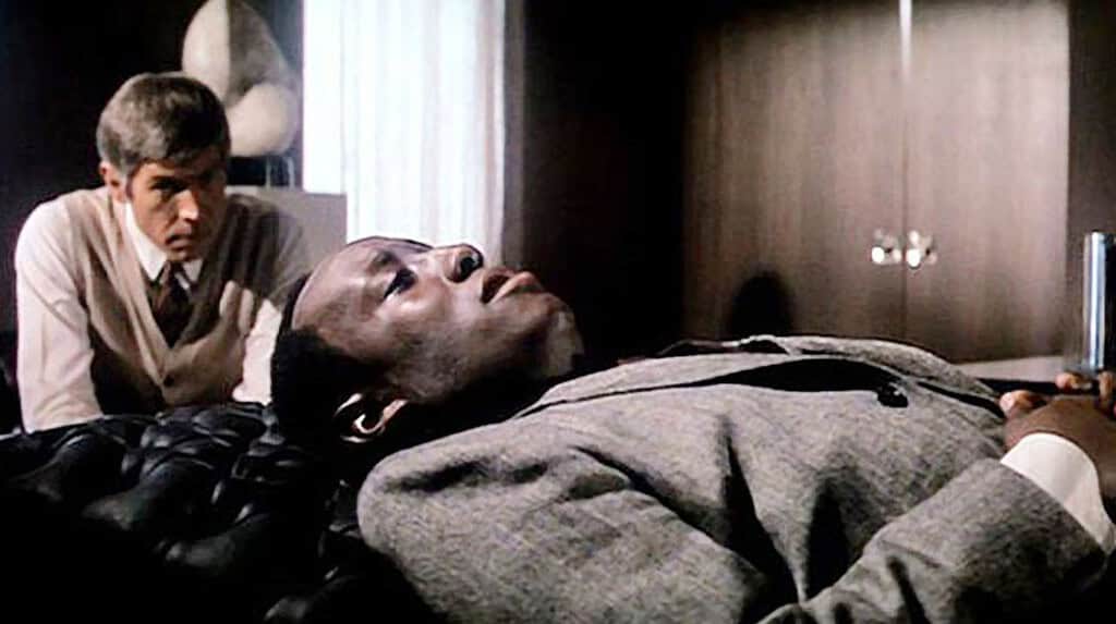 A scene from a film showing a white man in a sweater vest attending to a black man lying on a couch, looking upwards pensively.