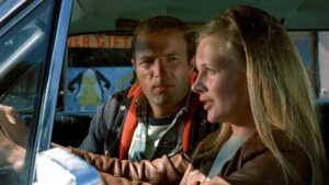 A man and woman are sitting in the front seats of a car. The woman is driving while talking, and the man is looking at her. A store sign is visible in the background through the window.