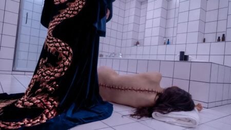 A person lies face-down on the floor of a tiled bathroom with a large stitched incision on their back. Another figure stands nearby wearing a dark robe with a gold dragon design.