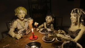 Three sculptures resembling people dine at a candlelit table, covered in textured materials and paint, evoking a surreal dinner scene.