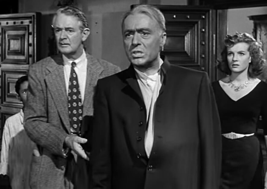 Black and white film scene featuring an older man in the foreground with two men and a woman looking on from the background.