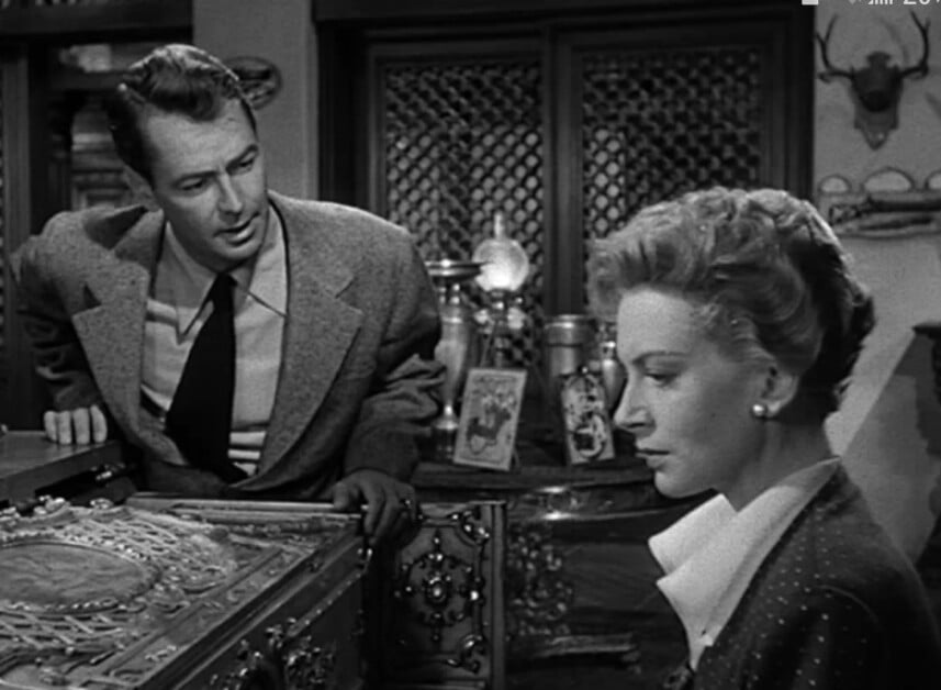 Two people, a man and a woman, in a vintage black and white film scene, having a conversation beside an ornate chest.