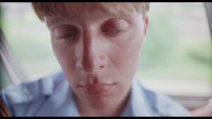 ‘Too Much Sleep’ Blu-Ray Review – Rediscovering A Lost Odyssey Close-up of a person with short hair and light skin, wearing a blue uniform shirt. The person appears to be looking down, with a blurred green background behind them.