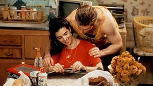 A man and woman sitting at a kitchen table, smiling and talking while looking at a photo album, with groceries and flowers nearby.