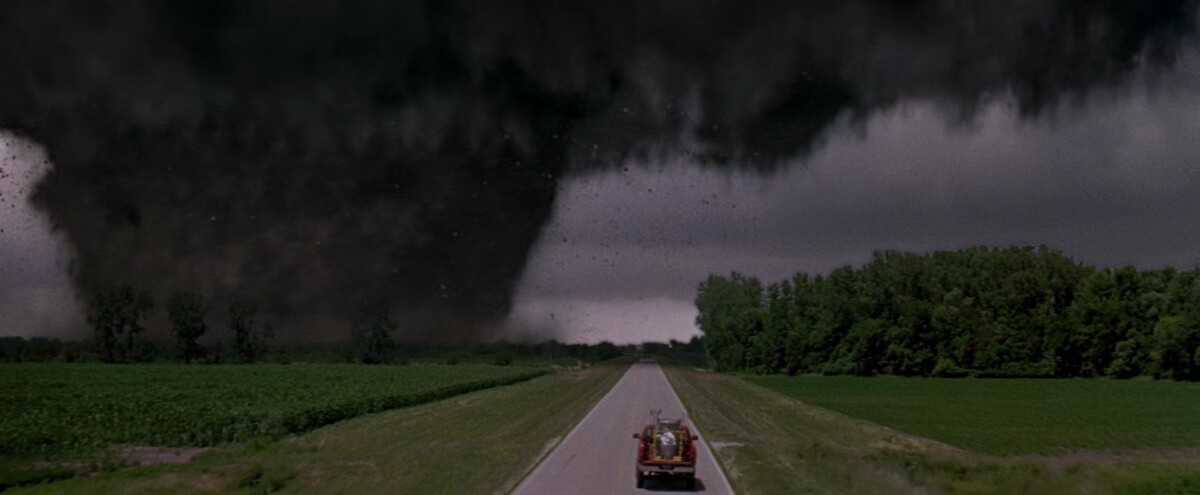 A large tornado approaches a vehicle driving on a rural road flanked by green fields and trees under a dark stormy sky.