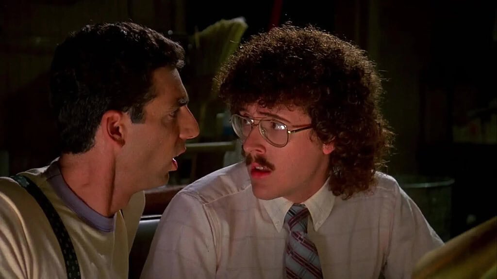 Two men in conversation; one is wearing a tank top and suspenders, the other glasses and a tie, in a dimly lit room.