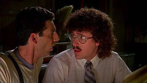 Two men in conversation; one is wearing a tank top and suspenders, the other glasses and a tie, in a dimly lit room.