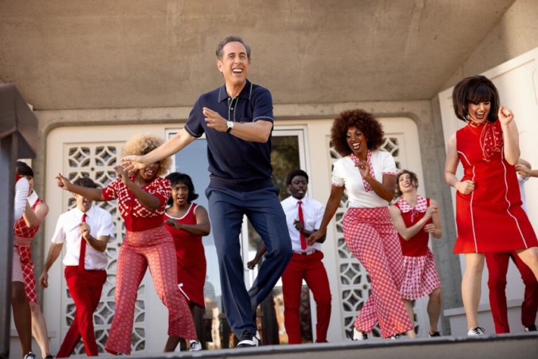 A man in a navy shirt and jeans dances with a group of people in red and white costumes on an outdoor stage.
