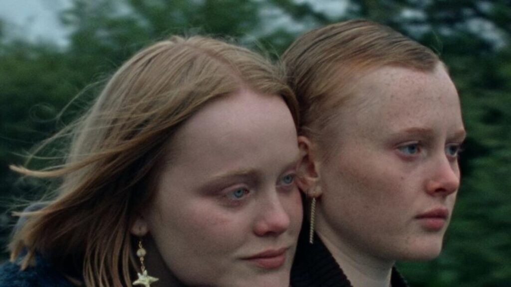 Two young individuals, one wearing star-shaped earrings, appear solemn and teary-eyed, standing outdoors with blurred greenery in the background.