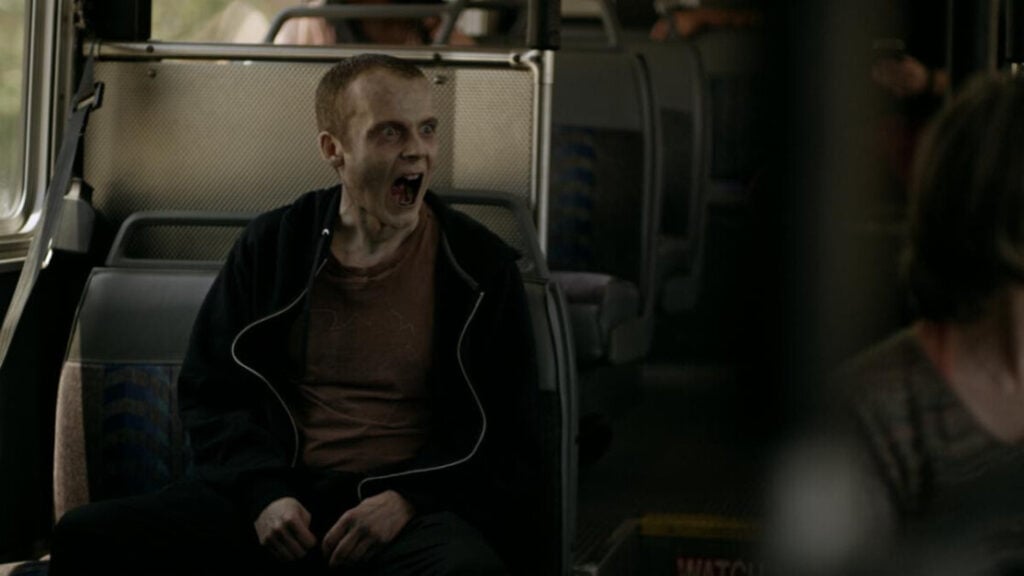 A person with pale skin and dark under-eye circles sits on a bus seat, mouth open wide as if shouting or yawning, wearing a dark hoodie and an orange shirt.