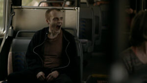 A person with pale skin and dark under-eye circles sits on a bus seat, mouth open wide as if shouting or yawning, wearing a dark hoodie and an orange shirt.