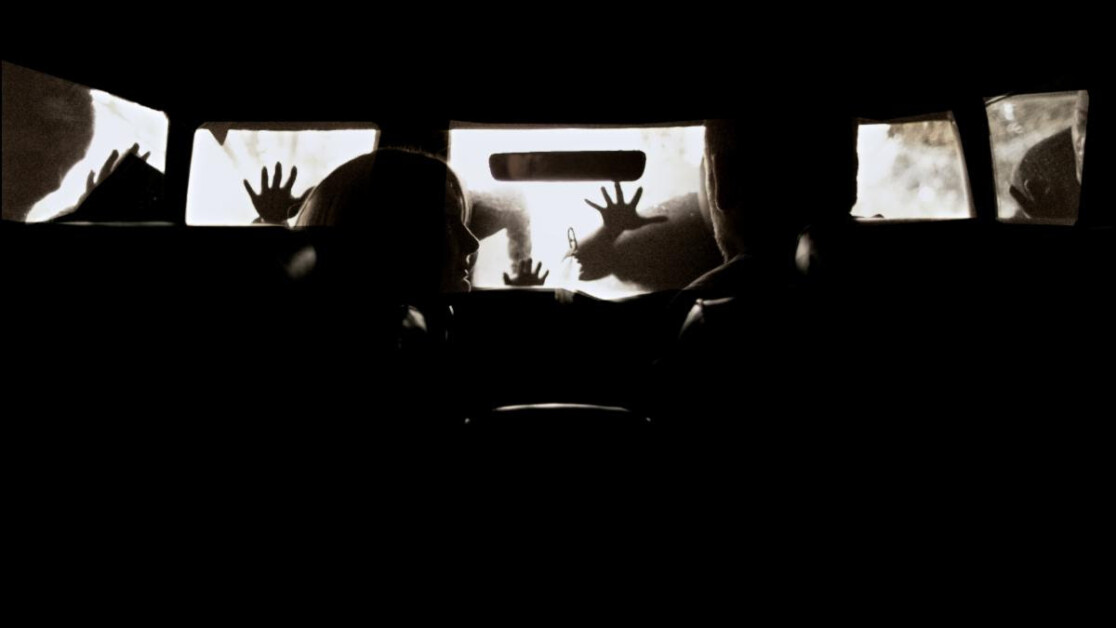 Silhouetted view of two people sitting in a car with several hands pressing against the car windows from outside. The scene appears tense and dramatic.