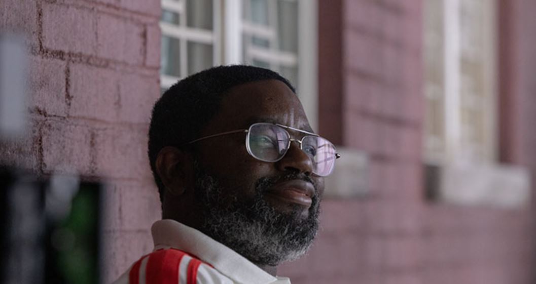 A man with glasses and a beard stands against a brick wall wearing a white shirt with red stripes.