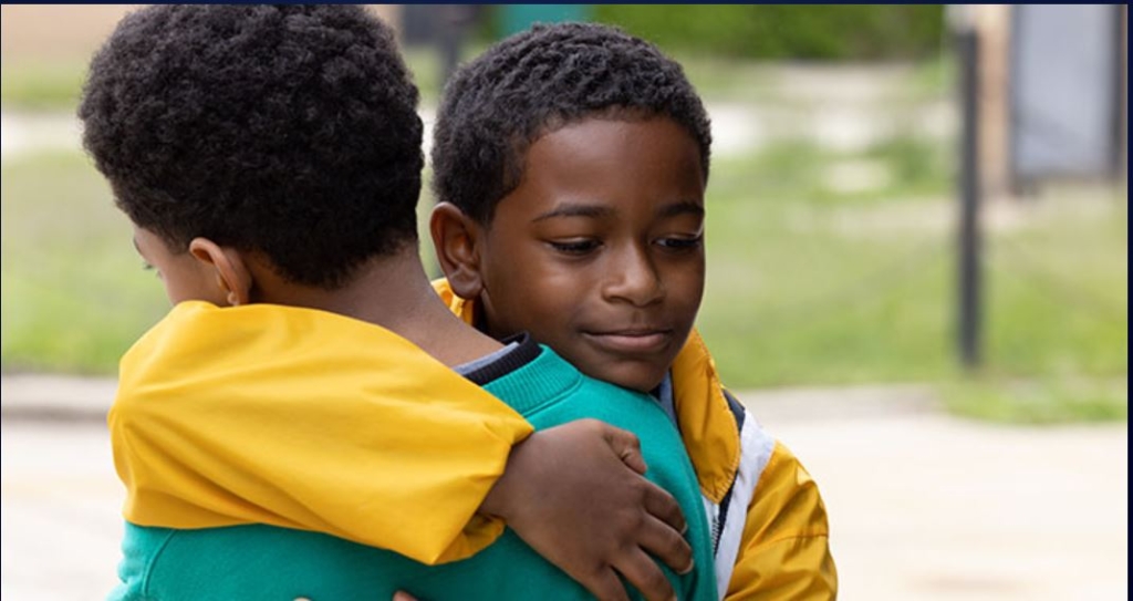 Two children with short curly hair hug each other outdoors. One child wears a yellow jacket and green shirt, while the other wears a green jacket.