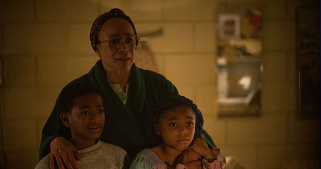 An older woman stands with her arms around two children, a boy and a girl wearing nightclothes, in a dimly lit room.