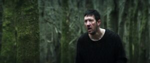 ‘Without Name’ Blu-Ray Review – Folk Horror Outing Conjures Lingering Dread A distressed man with short, dark hair and facial cuts is wearing a worn, dark sweater and standing in a dense, forested area. The trees in the background are covered with moss.