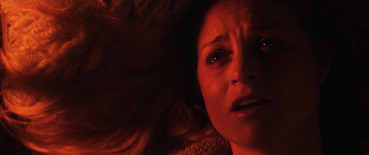 Close-up of a woman lying down with a concerned expression, illuminated by warm, orange-red lighting. Her head is partially on a textured surface, and her hair is spread out.