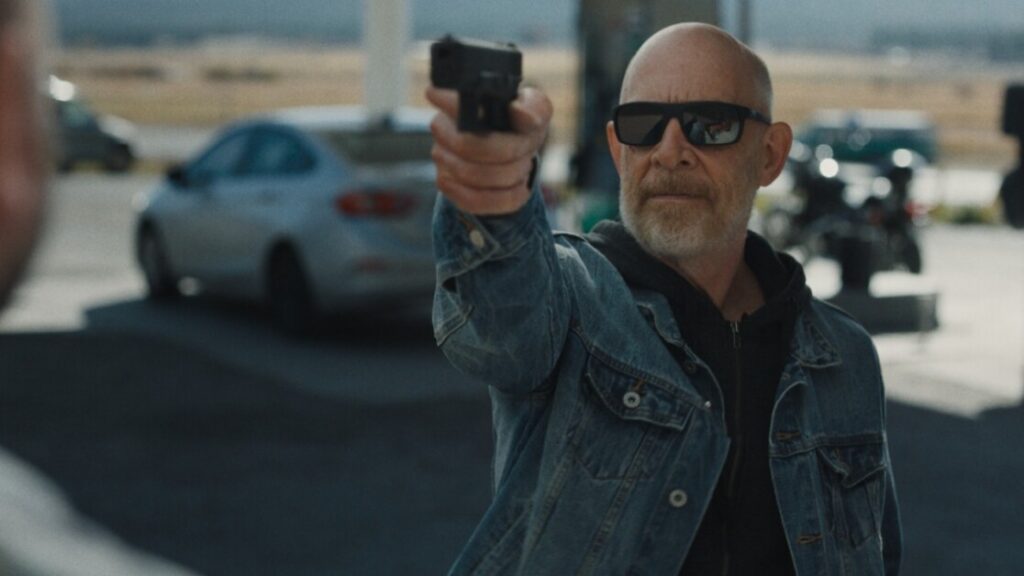 A bald man with glasses and a denim jacket points a handgun forward. There are cars and a motorcycle in the background, suggesting an outdoor or roadside setting.