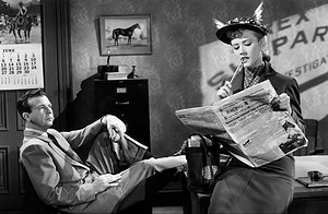 ‘You Never Can Tell’ Blu-Ray Review – More Peculiar Than You Could Ever Imagine A woman in a quirky hat reads the racing news in a newspaper while a man looks on, seated in an office-like room with a june calendar and racing posters on the wall.