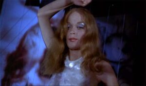 A person with long red hair and shimmering makeup stands with one arm raised against a blurred abstract background.