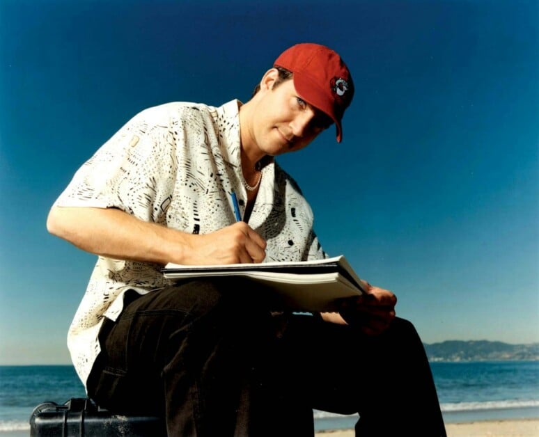A person wearing a red cap and patterned shirt is sitting outdoors, writing in a notebook with a beach and mountains in the background.