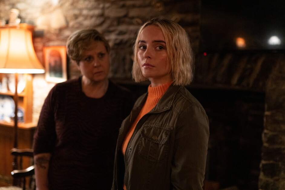 Two women stand in a dimly lit room with stone walls; the woman in front looks directly at the camera, while the woman behind has a concerned expression and looks at the first woman.