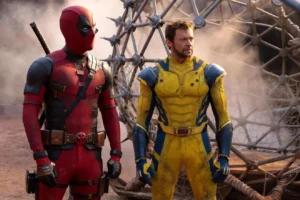 Two people in superhero costumes stand side by side. The person on the left is in a red and black suit with a mask covering their face, while the one on the right wears a yellow and blue suit.