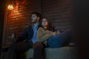 A man and a woman sit closely together on a bench, holding bottles and looking to the side. They appear to be in a dimly lit, rustic setting with wooden walls and a small light overhead.