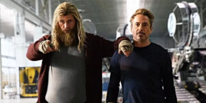 Two men stand side by side in an industrial setting. The man on the left has long blond hair and a bushy beard, wearing a cardigan. The man on the right has shorter brown hair and is wearing a dark top.