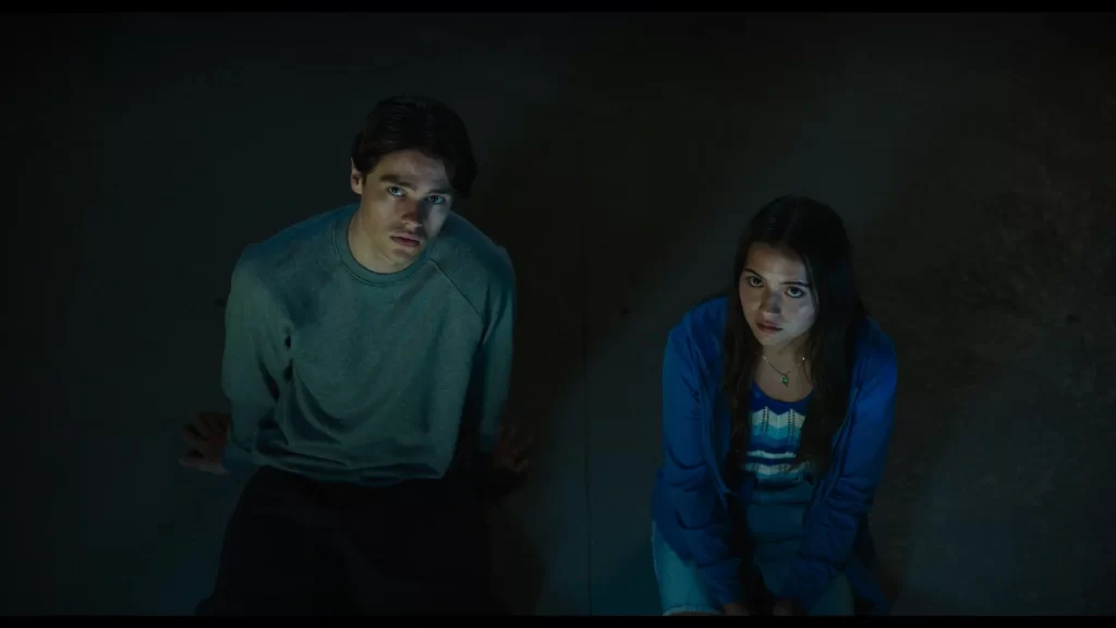 Two young adults, a man and a woman, sit on the ground in a dimly lit area, looking upwards with concerned expressions.