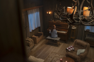 A person sits at a piano in a dimly lit rustic room with wooden walls, an antler chandelier, and cozy furniture.