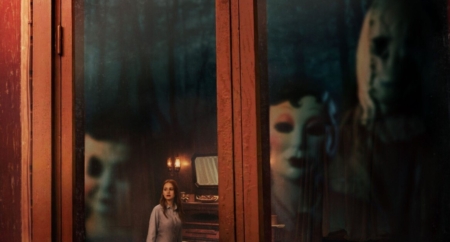 A person stands in a dimly lit room, looking through a glass door. Two figures in masks are visible behind the glass.