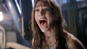 A woman with wet hair and a necklace screams with her mouth wide open in a dimly lit environment.