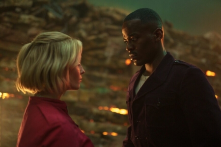 A close-up of a man and woman facing each other with intense expressions in a dimly lit setting with warm backlighting.